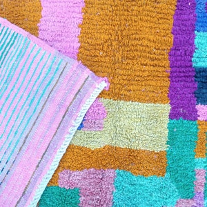 K&ouml;nnte beinhalten: Ein bunter Teppich mit einem geometrischen Muster in den Farben Rosa, Orange, Gelb, Gr&uuml;n, Blau und Lila. Der Teppich ist aus einem weichen, strukturierten Material gefertigt.