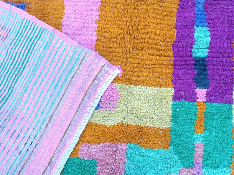 K&ouml;nnte beinhalten: Ein bunter Teppich mit einem geometrischen Muster in den Farben Rosa, Orange, Gelb, Gr&uuml;n, Blau und Lila. Der Teppich ist aus einem weichen, strukturierten Material gefertigt.