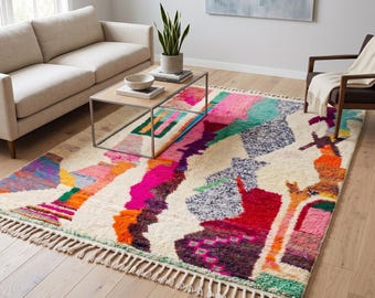 May include: A rectangular area rug featuring a cream-colored base with an abstract, multi-colored pattern in shades of hot pink, orange, teal, purple, and green. The rug has a thick, textured pile and is finished with knotted fringe on both ends.