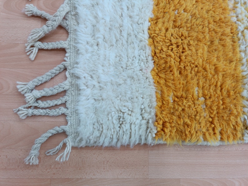 May include: A white and yellow woven rug with fringe on the edges. The rug has a simple design with two colors.