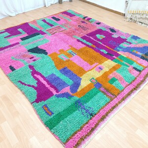 K&ouml;nnte beinhalten: Ein bunter marokkanischer Teppich mit einem geometrischen Muster in den Farben Rosa, Gr&uuml;n, Lila, Orange und Gelb. Der Teppich hat einen Fransenrand und ist aus Wolle gefertigt.