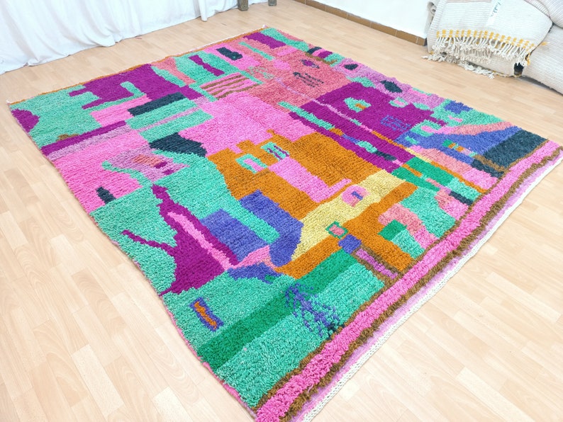 K&ouml;nnte beinhalten: Ein bunter marokkanischer Teppich mit einem geometrischen Muster in den Farben Rosa, Gr&uuml;n, Lila, Orange und Gelb. Der Teppich hat einen Fransenrand und ist aus Wolle gefertigt.