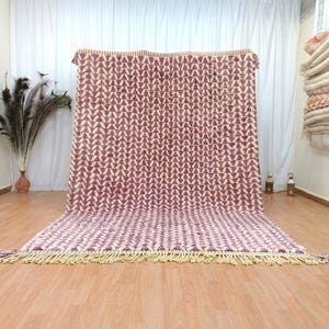 May include: A large, rectangular rug with a repeating chevron pattern in shades of burgundy and cream. The rug has a fringe along the bottom edge. The rug is displayed indoors, with a wooden floor and white curtains in the background.