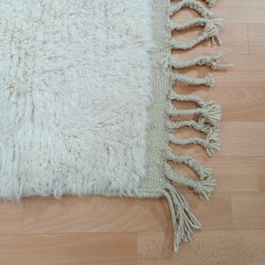 May include: A white fluffy rug with a fringe border. The rug is on a light wood floor.