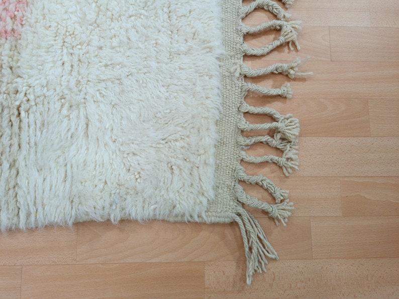May include: A white fluffy rug with a fringe border. The rug is on a light wood floor.