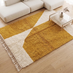 Puede incluir: Una alfombra rectangular de pelo largo con un patrón geométrico en color amarillo mostaza y blanco crema. La alfombra incluye detalles de flecos decorativos a lo largo de los bordes más cortos, creando una superficie suave y texturizada.