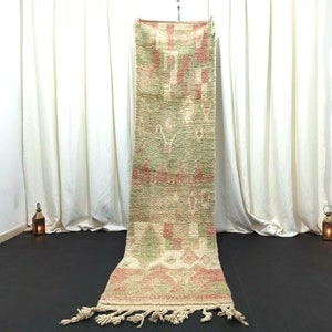 May include: A long, rectangular rug with a cream, green, and pink geometric pattern. The rug has fringe on the ends and is hanging from a rod.