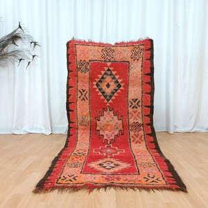 May include: A red and brown Moroccan rug with intricate geometric patterns. The rug has a fringed edge and is made of wool.