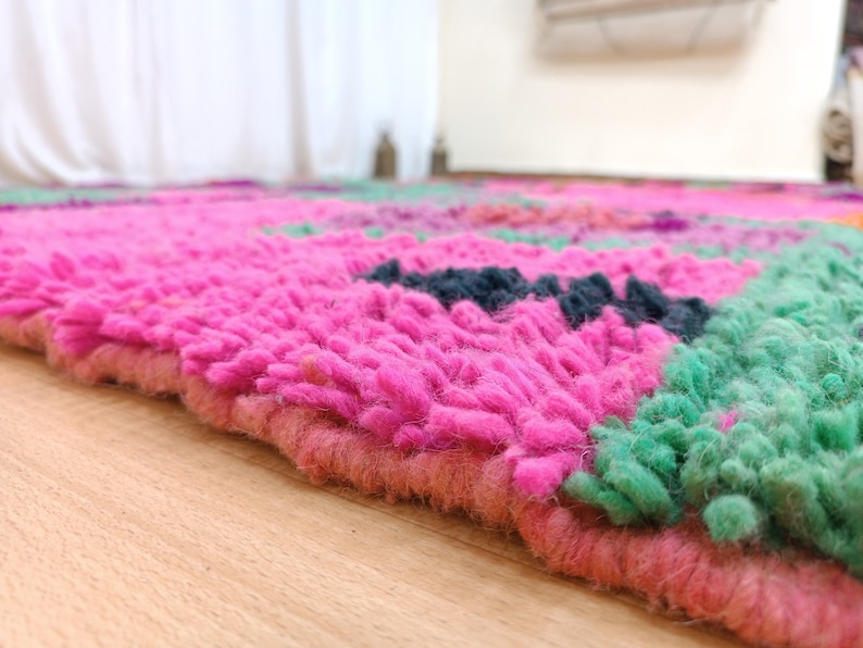 K&ouml;nnte beinhalten: Nahaufnahme eines handgewebten rosa und gr&uuml;nen Teppichs mit einer flauschigen Textur. Der Teppich hat ein geometrisches Muster mit Quadraten und Rechtecken.