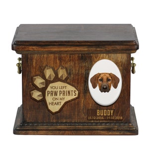 May include: Wooden pet urn with a paw print design and the text "You left paw prints on my heart". A portrait of a brown dog is on the right side of the urn. The text "Buddy 15.12.2005 - 19.07.2018" is below the portrait.