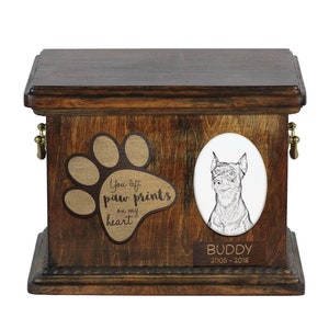 Könnte beinhalten: Eine hölzerne Tierurne mit einem Pfotenabdruck-Design und dem Text "You left paw prints on my heart". Ein Porträt eines Hundes befindet sich in einem weißen ovalen Rahmen. Die Urne hat eine Messing-Namenschild mit dem Namen "Buddy" und den Daten "2005-2018".