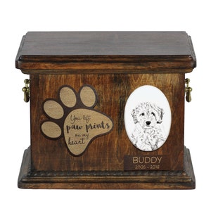 May include: Wooden pet urn with a paw print design and the text "You left paw prints on my heart". A white oval with a black and white drawing of a dog is on the front of the urn. The name "BUDDY" and the dates "2005-2018" are engraved on a plaque below the oval.