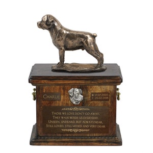 May include: A wooden pet urn with a bronze figurine of a Rottweiler dog on top. The urn has a plaque with the name "Charlie" and dates "17.07.2005" and "25.02.2018". The plaque also has the inscription "Those we love don't go away, they walk beside us everyday, unseen, unheard, but always near, still loved, still missed and very dear."