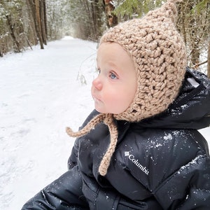 May include: A baby wearing a black snowsuit with a Columbia logo and a beige crocheted hat with a pom pom on top. The baby is sitting in the snow and looking to the right.