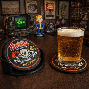 May include: A set of black RobCo Industries coasters with a robot graphic and the text "BUILT TO LAST!" A glass of beer sits on one coaster. The coasters are in a black holder on a wooden surface. The image has a retro video game theme.