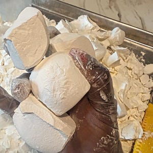 May include: A hand wearing a clear plastic glove holds several large, irregular, matte white chalk chunks. The chalk pieces have a porous, textured surface and are piled together with smaller fragments on a metallic tray with a yellow tool.