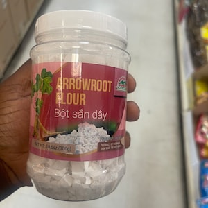 May include: A clear plastic jar of arrowroot flour, labeled with the text "ARROWROOT FLOUR" and "Bột sắn dây". The jar contains a white powder and has a pink and white label. The net weight is 10.5oz (300g).