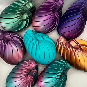 May include: A collection of shell-shaped objects in various colours, including green, purple, teal, and gold. Each object has a ribbed texture and a decorative element at the top. The objects appear to be decorative or artistic in nature.