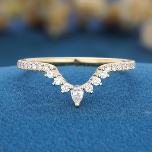 May include: A gold V-shaped ring featuring a series of small round-cut white stones along the band, culminating in a single pear-cut white stone at the center point. The band has a polished finish and follows a curved, chevron-like silhouette.