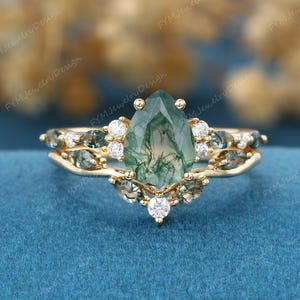 May include: A gold ring set featuring a pear-shaped moss agate center stone with green inclusions, accented by smaller marquise-cut moss agate stones and round white stones. The band has a curved design that follows the shape of the center stone.