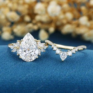 May include: A gold engagement ring set featuring a large pear-cut center stone flanked by marquise-cut side stones, paired with a curved wedding band adorned with round-cut stones that follow the contour of the pear-shaped center diamond.