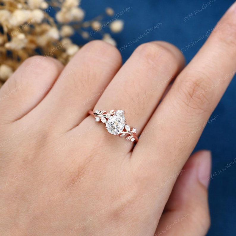 Op de afbeelding: Rosegouden verlovingsring met een peervormige diamant als middensteen. De band is versierd met delicate bladeren en kleine diamantaccenten. De ring wordt getoond op een hand tegen een blauwe achtergrond.