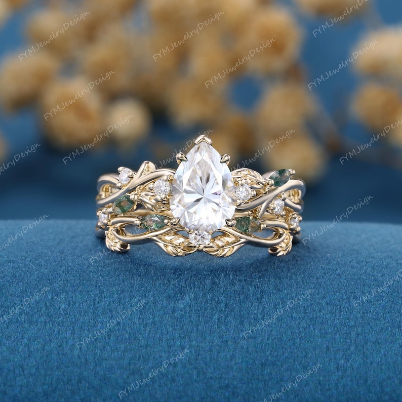 May include: A three-piece gold ring set. The engagement ring features a pear-shaped diamond. The band and the other ring have a leaf design with small diamonds and green gemstones. The rings are on a blue surface.