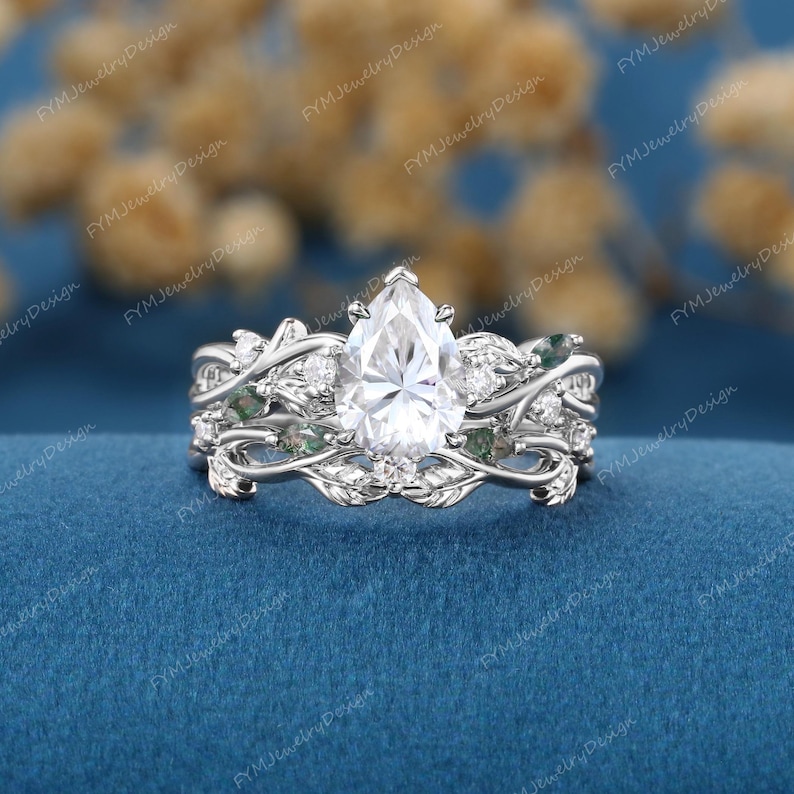 May include: A three-piece bridal ring set featuring a pear-shaped, clear gemstone center stone. The rings have a floral vine design with small accent stones and green accents. The rings are set on a blue surface.
