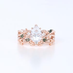 May include: Rose gold engagement ring set featuring a pear-shaped center stone. The band is adorned with leaf and vine details, small clear stones, and green accents. The set includes a matching band with similar design elements.