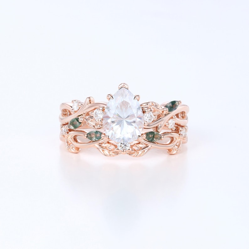 May include: Rose gold engagement ring set featuring a pear-shaped center stone. The band is adorned with leaf and vine details, small clear stones, and green accents. The set includes a matching band with similar design elements.