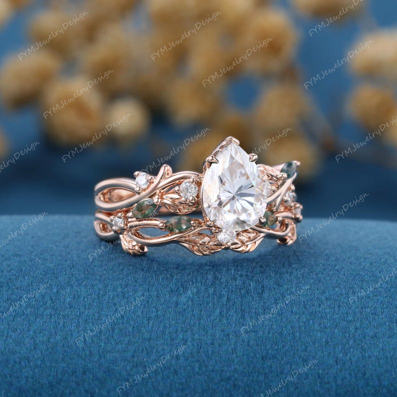 May include: Rose gold engagement ring set featuring a pear-shaped, clear gemstone. The rings have a nature-inspired design with leaf and vine details, accented with small, round gemstones and green stones. The set is displayed on a blue surface.