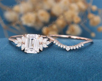 May include: A rose gold engagement ring set featuring a central emerald-cut stone flanked by marquise-cut stones, paired with a curved wedding band adorned with a row of small round-cut stones, both set on a thin, polished rose gold metal band.