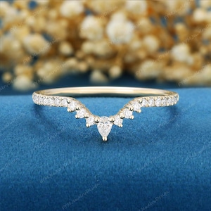 May include: A gold chevron-shaped ring featuring a series of small round-cut clear stones along the band, culminating in a single pear-cut clear stone at the center point. The band has a polished finish and follows a V-shaped contour design.