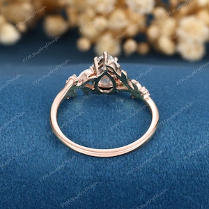 Op de afbeelding: Rosegouden verlovingsring met een peervormige centrale steen en bladachtige accenten. De ring heeft een delicate band en is geplaatst tegen een blauwe achtergrond. Het ontwerp van de ring omvat kleinere stenen aan weerszijden van de hoofdsteen.