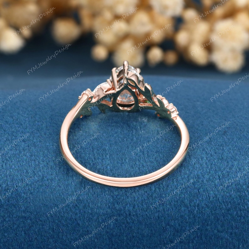 Op de afbeelding: Rosegouden verlovingsring met een peervormige centrale steen en bladachtige accenten. De ring heeft een delicate band en is geplaatst tegen een blauwe achtergrond. Het ontwerp van de ring omvat kleinere stenen aan weerszijden van de hoofdsteen.