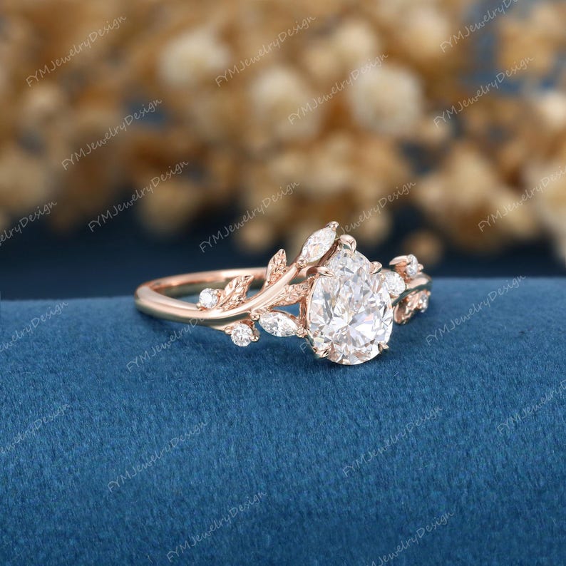 Op de afbeelding: Rosegouden verlovingsring met een peervormige diamant als middensteen. De band heeft een delicaat blad- en marquise-diamanten ontwerp. De ring is geplaatst tegen een blauwe achtergrond.
