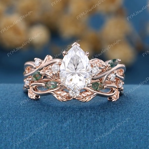 Peut inclure: Une bague en or rose ornée d'une pierre centrale transparente taille poire, entourée de petites pierres transparentes rondes et de pierres vertes effet mousse taille marquise. L'anneau présente des motifs de feuilles et de vignes texturés.