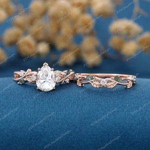May include: Rose gold engagement ring set with a pear-shaped center stone and a matching band. The rings feature leaf and vine detailing with small accent stones and green accents. The background is a deep blue.