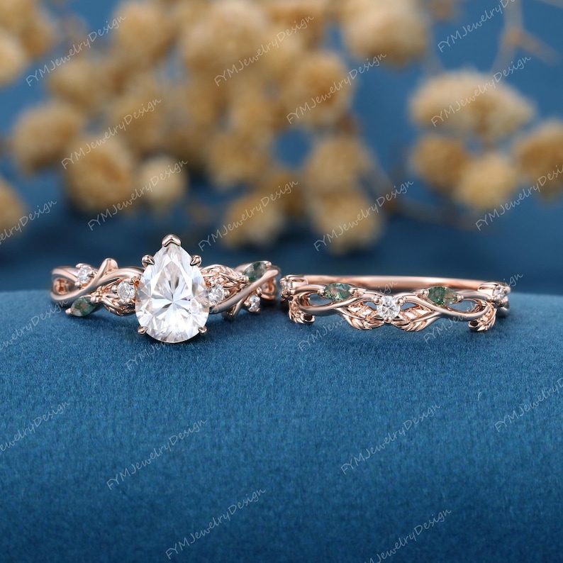 May include: Rose gold engagement ring set with a pear-shaped center stone and a matching band. The rings feature leaf and vine detailing with small accent stones and green accents. The background is a deep blue.