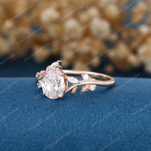 Op de afbeelding: Een ros&eacute;gouden verlovingsring met een peervormige diamant als middensteen. De ring heeft delicate blad- en marquise-geslepen diamantaccenten. De ring ligt op een blauw oppervlak.