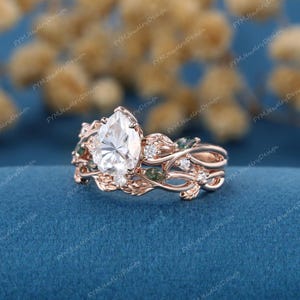 May include: Rose gold engagement ring set with a pear-shaped center stone and leaf-like detailing. The ring features small accent stones and green gemstones, creating a nature-inspired design. The ring is set against a blue background.
