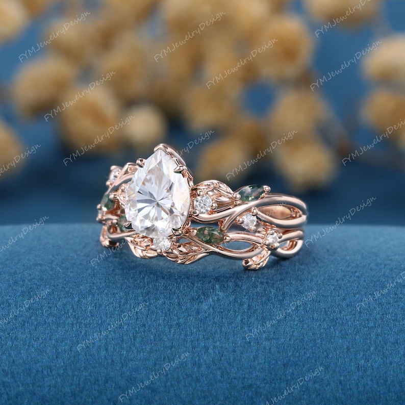 May include: Rose gold engagement ring set with a pear-shaped center stone and leaf-like detailing. The ring features small accent stones and green gemstones, creating a nature-inspired design. The ring is set against a blue background.