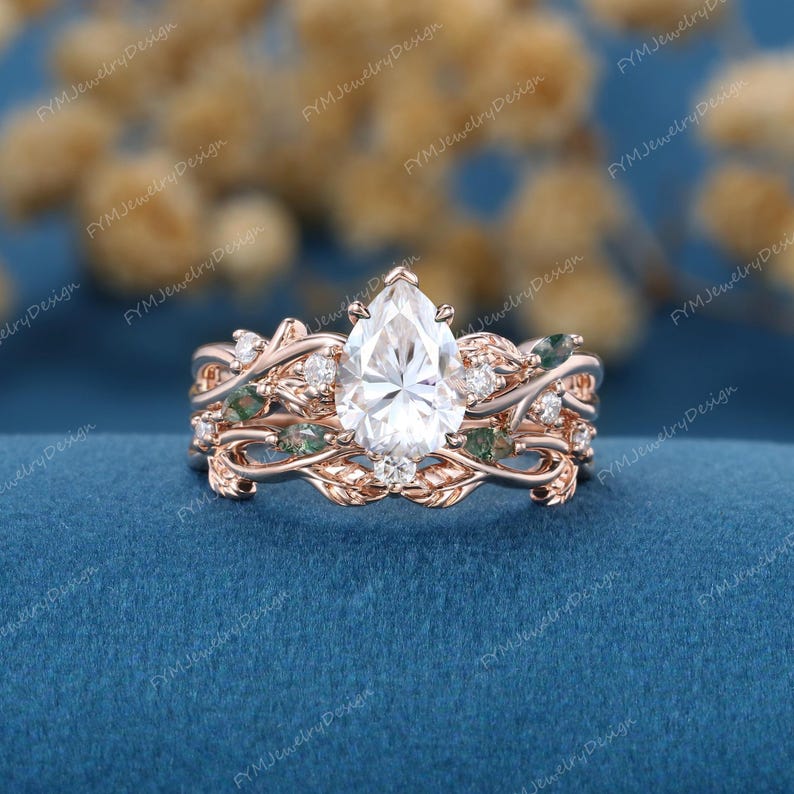 May include: A rose gold engagement ring set featuring a pear-shaped center stone, surrounded by smaller diamonds and green accents. The band has a nature-inspired design with leaf and vine details. The ring set is displayed on a blue surface.