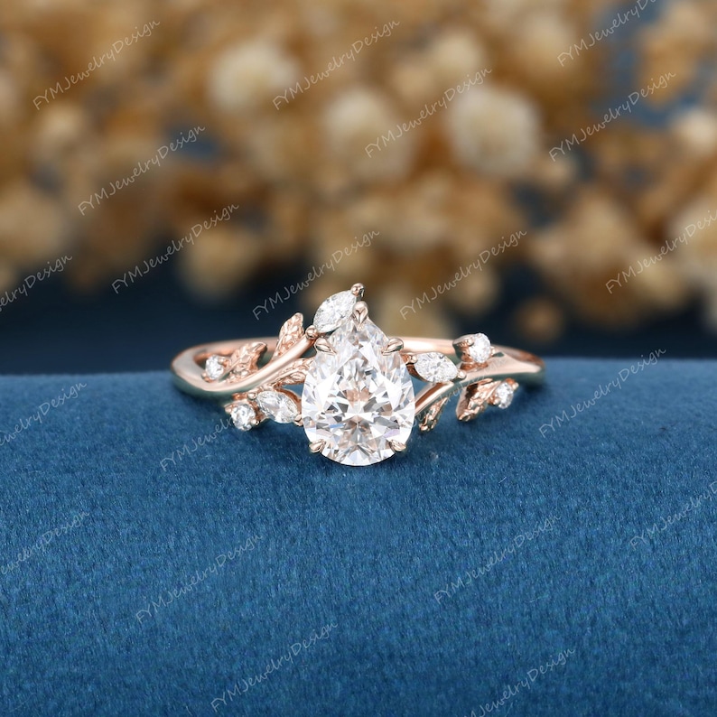 May include: Rose gold engagement ring featuring a pear-shaped diamond center stone. The ring has a delicate vine and leaf design with smaller accent diamonds. The ring is set on a blue background.