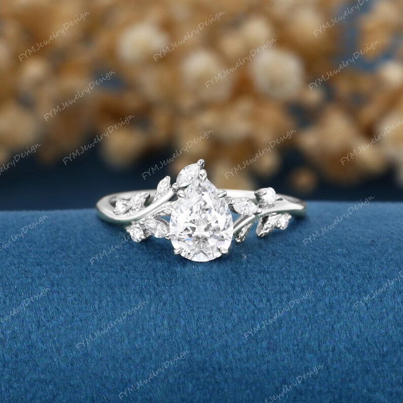 Op de afbeelding: Een zilveren ring met een peervormige, heldere edelsteen als middelpunt. De ring heeft een bladeren ontwerp met kleinere, ronde edelstenen. De ring is geplaatst tegen een blauwe achtergrond.
