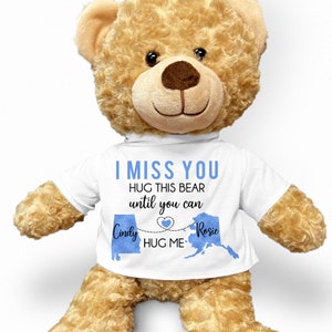 May include: A brown teddy bear wearing a white t-shirt with the text "I miss you. Hug this bear until you can." and a blue heart with a line connecting two blue states with the text "Cindy" and "Rosie" and "Hug me".