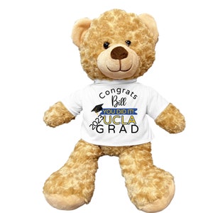 Puede incluir: Un oso de peluche marr&oacute;n que lleva una camiseta blanca con el texto "Congrats Bill You Did It! 2023 UCLA GRAD" en letras negras y azules.