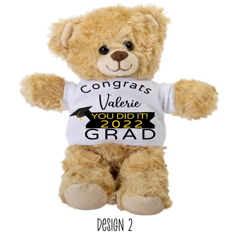 Puede incluir: Un oso de peluche marr&oacute;n que lleva una camiseta blanca con el texto "Congrats Valerie You Did It! 2022 Grad" en negro y amarillo. La camiseta tiene un gr&aacute;fico de gorro de graduaci&oacute;n negro.