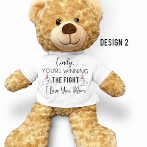 May include: A brown teddy bear wearing a white t-shirt with the text "Cindy, You're Winning The Fight I Love You, Mom" and pink ribbon boxing gloves. The text "DESIGN 2" is in the upper right corner of the image.