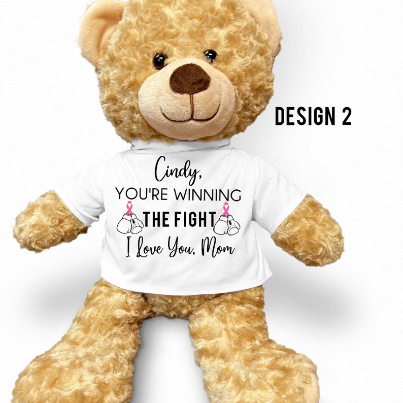 May include: A brown teddy bear wearing a white t-shirt with the text "Cindy, You're Winning The Fight I Love You, Mom" and pink ribbon boxing gloves. The text "DESIGN 2" is in the upper right corner of the image.